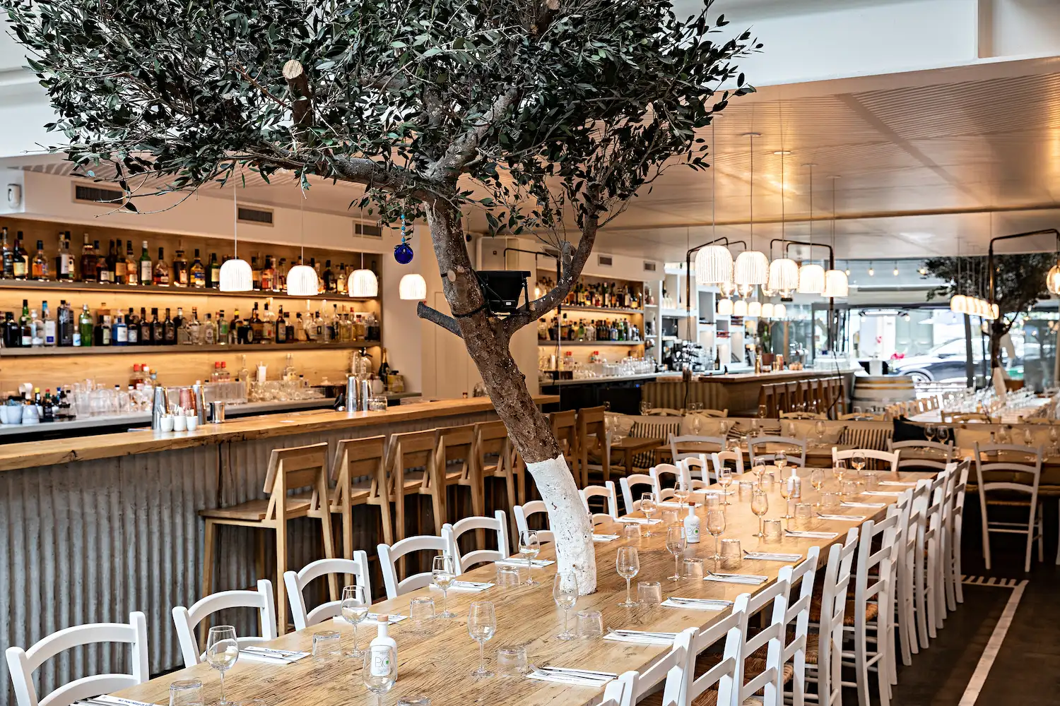 Warm event restaurant with long tables, central bar and indoor olive trees